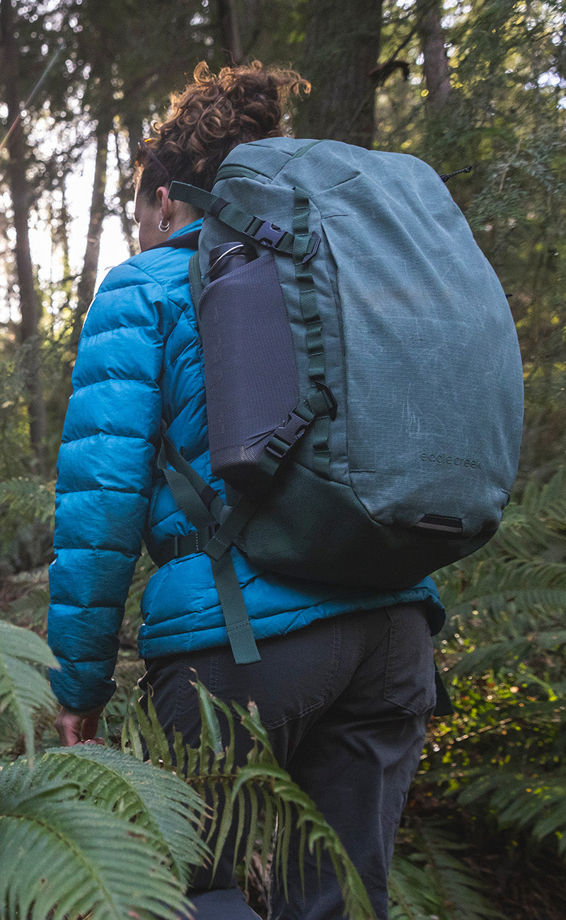 A hiker walks through a lush forest wearing a green Eagle Creek backpack.