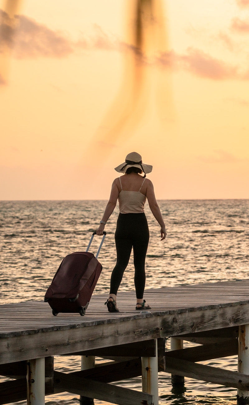 A woman walking on a wooden pier by the ocean at sunset, pulling a carry-style travel luggage bag with small wheels.