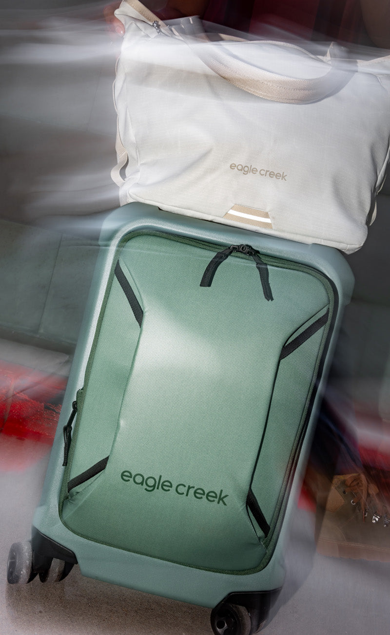 A close up image of a green rolling luggage bag and a white tote bag for travel on top of it.
