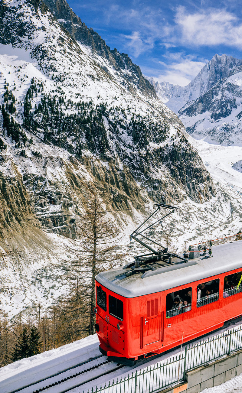 A red train passes through a sunny snowy mountainous area.
