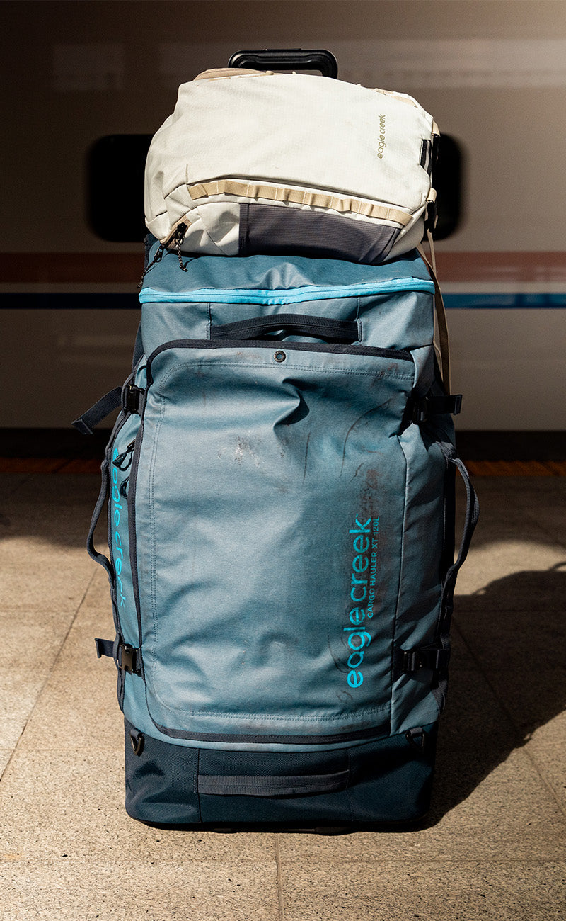 A white travel backpack sits on top of a large blue rolling duffle bag.